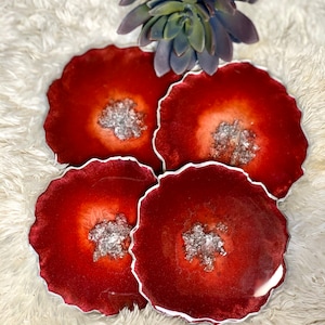 May include: Four red and silver resin coasters with a wavy edge. The coasters have a white border and a silver glitter design in the center.