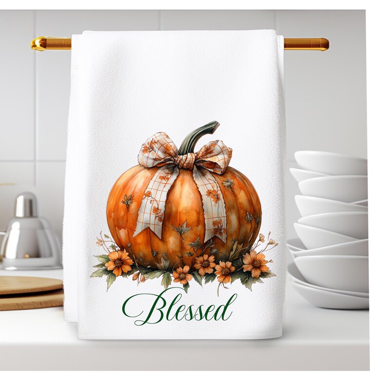 Op de afbeelding: Witte keukendoek met een handgeschilderde illustratie van een oranje pompoen met een geruite strik en groene bladeren. De pompoen is omringd door kleine gele bloemen. De tekst "Blessed" is in groene cursieve letters onder de pompoen geschreven.