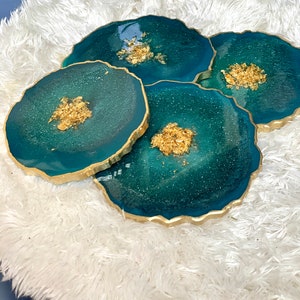 May include: Four teal blue and gold resin coasters with a glittery finish. The coasters have a unique, irregular shape and are perfect for adding a touch of elegance to any home.