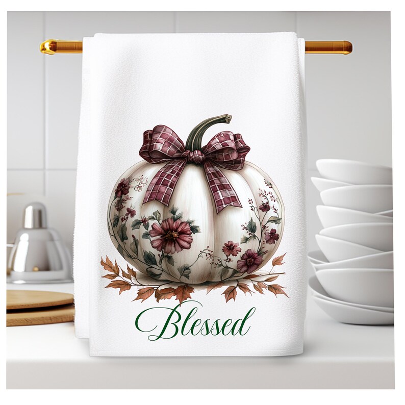 Op de afbeelding: Witte pompoen met een rood-wit geruite strik en bloemdetails. De pompoen is omringd door herfstbladeren en het woord "Blessed" staat eronder.