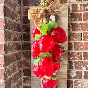 May include: A wooden sign with a cluster of bright red strawberries and green leaves. It has a burlap bow and a natural rope for hanging. The background is a rustic wooden plank, ideal for a kitchen or dining area.