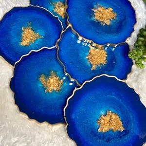 May include: Set of five blue and gold resin coasters with a unique, irregular shape. Each coaster features a swirling blue design with gold flecks.