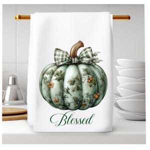 Op de afbeelding: Een witte keukendoek met een groene pompoen met een geruite strik en groene bladeren. De pompoen heeft een bloemenpatroon en de tekst "Blessed" is onder de pompoen gedrukt.