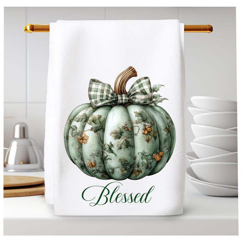 Op de afbeelding: Een witte keukendoek met een groene pompoen met een geruite strik en groene bladeren. De pompoen heeft een bloemenpatroon en de tekst "Blessed" is onder de pompoen gedrukt.