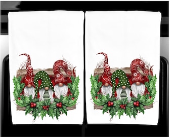 Puede incluir: Dos paños de cocina blancos con un diseño festivo de gnomos. Cada paño presenta tres gnomos con sombreros rojos estampados, sombreros verdes con lunares blancos y barbas blancas, rodeados de acebo y bayas.