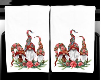Puede incluir: Dos paños de cocina blancos con un diseño de gnomo navideño. Cada paño presenta tres gnomos con sombreros rojos y estampados florales y barbas blancas. Los gnomos están rodeados por una corona de vegetación y poinsettias rojas.