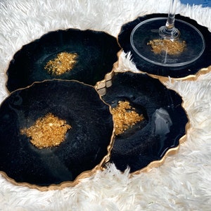 May include: Set of four black and gold resin coasters with a wavy edge. Each coaster has a gold fleck design.