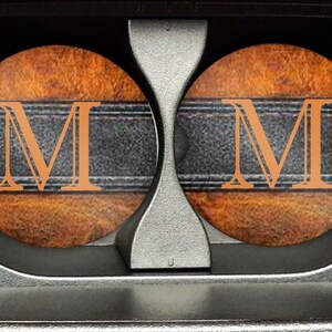 May include: Two brown and orange coasters with a black leather-like band and the letter "M" in orange. The coasters are in a car cup holder.
