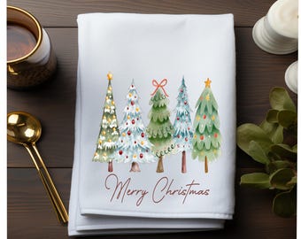 May include: White Christmas-themed kitchen towel featuring a watercolor illustration of five decorated Christmas trees. The words "Merry Christmas" are written in a cursive font below the trees. The towel is folded and placed on a dark wooden surface.