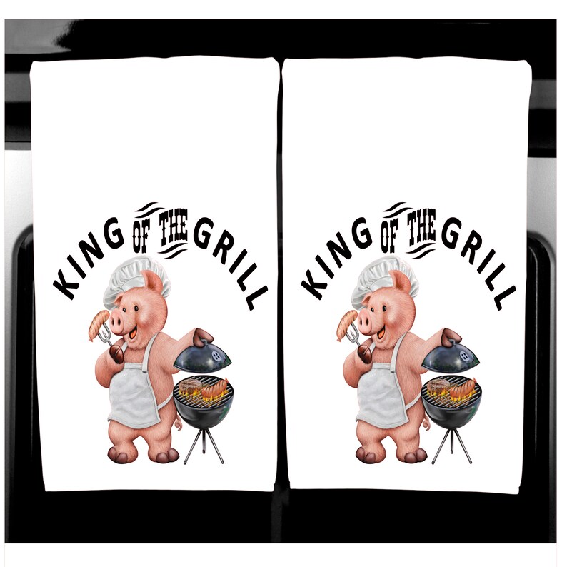 K&ouml;nnte beinhalten: Zwei wei&szlig;e K&uuml;chent&uuml;cher mit einem Cartoon-Schwein-Koch und einem Grill. Die T&uuml;cher zeigen den Text "KING OF THE GRILL" in Schwarz, der &uuml;ber der Schweine-Illustration angeordnet ist. Das Schwein tr&auml;gt eine Kochm&uuml;tze und eine Sch&uuml;rze.