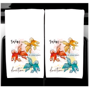 K&ouml;nnte beinhalten: Zwei wei&szlig;e K&uuml;chent&uuml;cher mit einem Aquarelldesign von Goldfischen in Orange, Gelb und T&uuml;rkis. Die T&uuml;cher tragen den Schriftzug "swim in your own direction" in schwarzer Schrift.