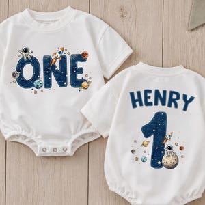 May include: Two white short-sleeved baby bodysuits featuring space-themed graphics. One bodysuit displays the word ONE in dark blue with stars, planets, a rocket, and an astronaut. The other displays the name HENRY above the number 1.