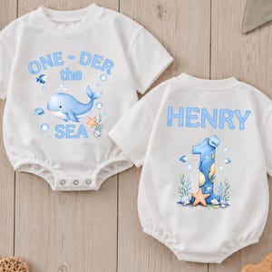 May include: Two white short-sleeved baby bodysuits featuring blue graphics. One bodysuit displays a whale and the text ONE-DER the SEA. The other bodysuit features the name HENRY above a large number 1 decorated with seashells and a starfish.