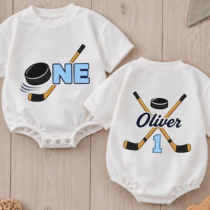 May include: Two white short-sleeved baby bodysuits featuring hockey-themed graphics. One displays a hockey stick, a puck, and the word ONE in blue. The other shows two crossed hockey sticks, a puck, the name Oliver, and the number 1 in blue.