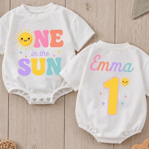 May include: Two white short-sleeved baby bodysuits. The left one features a yellow sun graphic and the text "ONE in the SUN" in pink, orange, yellow, and blue. The right one displays the name "Emma" and the number "1" with a small sun graphic.
