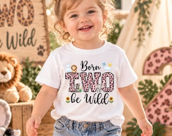 May include: A white short-sleeved t-shirt featuring a graphic print with the text Born TWO be Wild. The design includes leopard print lettering, small illustrations of a lion, zebra, elephant, and sunflowers, and a pink hair bow accessory.