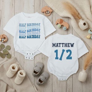 May include: Two white baby onesies with blue text. One says "HALF BIRTHDAY" multiple times, the other says "MATTHEW 1/2". The image includes baby shoes, socks, and wooden toys on a light wood surface.