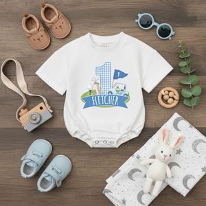 May include: A white baby onesie with a golf-themed design, including a blue gingham number one, golf cart, and the name "Fletcher" on a banner. The image also features baby shoes, sunglasses, a toy camera, and a plush bunny.