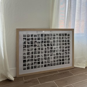 May include: A framed collage of numerous small, square black and white photographs. The frame is light-coloured wood, and the photos are arranged in a grid pattern. The artwork is displayed against a white wall, with sheer curtains visible in the background.