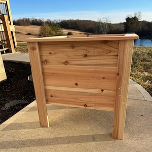 Peut inclure: Un bac à plantes rectangulaire en bois avec une finition naturelle. Le bac est construit à partir de planches horizontales et présente un design de pieds robuste et incliné. Le bac est vide et prêt à planter. Le fond montre un cadre extérieur naturel.