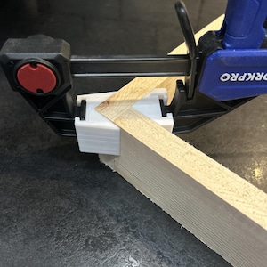 Puede incluir: Una abrazadera WORKPRO negra y azul asegura dos piezas de madera clara en un ángulo de 90 grados. La abrazadera, con un botón de liberación rojo, se utiliza para un proyecto de carpintería.