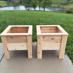 May include: Two wooden planters with a natural finish. Each planter has a square top and is constructed with light-colored wood. The planters are set on four legs and are suitable for outdoor use. The background features a pond and green grass.