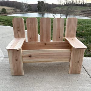 Könnte beinhalten: Eine Holzbank im Adirondack-Stil mit natürlicher Oberfläche. Die Bank hat eine Lattenlehne und Armlehnen. Die Bank ist aus hellem Holz gefertigt und für den Außenbereich konzipiert.