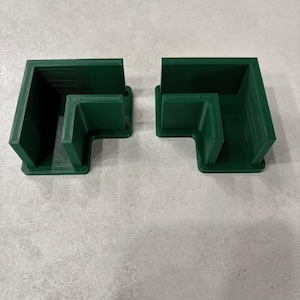 Peut inclure: Deux équerres d'angle imprimées en 3D, en forme de L, de couleur vert foncé. Chaque équerre a une conception à angle droit avec une base plate et des côtés verticaux. Les équerres sont en matériau opaque et sont posées sur une surface grise claire.