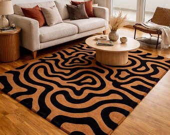 May include: A living room scene featuring a large area rug with a black and brown abstract design. The rug is the focal point, complemented by a beige sofa with decorative pillows, a wooden coffee table, and a wicker chair.