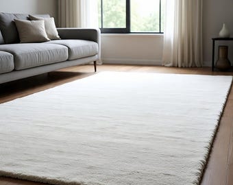 May include: A large, rectangular, off-white rug with a soft, textured surface. The rug is placed on a hardwood floor in a living room setting, with a gray sofa and window in the background. The rug's dimensions are not visible.