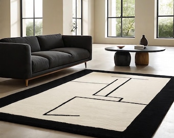 May include: A modern area rug with a cream-colored center and a bold black border. The rug features a geometric design in black lines, adding a contemporary touch to the living room setting. The rug is placed in front of a dark gray sofa.