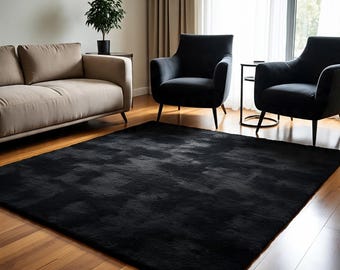 May include: A large, rectangular black rug with a textured surface dominates the foreground. The rug sits on a hardwood floor, and is surrounded by furniture, including a beige sofa and two black armchairs. A small potted plant is visible.
