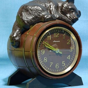 May include: A brown bear figurine rests atop a brown barrel-shaped clock. The clock face is brown with gold-colored hands and numerals. The clock has a black base and the word "Маяк" is visible on the clock face.