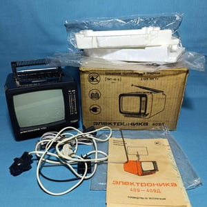 May include: A black portable Elektronika 409D television set with a handle, power cables, and a telescopic antenna. The set includes a cardboard box with white styrofoam packaging and a printed instruction manual featuring the model name 409-409D.