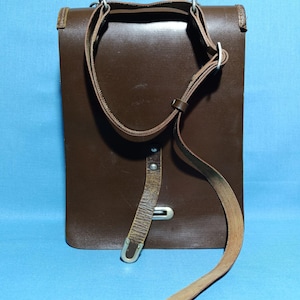 May include: A dark brown leather bag featuring a rectangular structure, a long adjustable shoulder strap, and a vertical leather closure strap secured with a silver-toned metal buckle. The bag has a smooth finish and visible metal hardware.