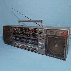 May include: A grey Sharp portable stereo system featuring dual cassette decks, a radio tuner with a long extendable antenna, and two large speakers with black metal mesh grilles. The front panel displays the text High-Speed Dubbing and SHARP.