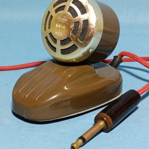 May include: Vintage brown microphone with a gold-colored mesh grill and a matching base. The microphone has a red cord and a brown plug. The text "MA-41" and "1960" are visible on the grill. The microphone is likely from the mid-20th century.