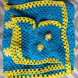 May include: A handmade crochet blanket in shades of blue and yellow. The blanket features a square pattern with a yellow border and decorative yellow floral accents. The blanket is folded, showcasing its texture and design.