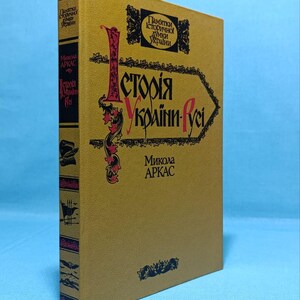 May include: A hardcover book with a gold cover and spine. The title "Історія України-Руси" is displayed in red and black lettering. The author's name, Микола Аркас, is also visible. The book has decorative elements and text in Ukrainian.