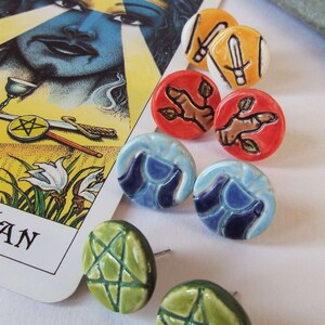 May include: A set of ceramic earrings featuring various designs, including a pentagram, swords, and a hand with leaves. The earrings are displayed on a white surface with a tarot card in the background.