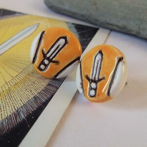 May include: A pair of orange and white ceramic earrings with a black sword design.