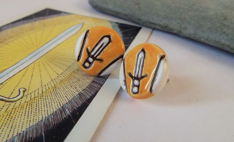 May include: A pair of orange and white ceramic earrings with a black sword design.