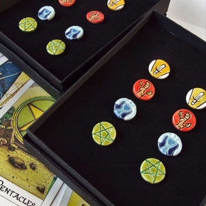 May include: Two black velvet boxes with a variety of colorful round ceramic tokens. The tokens feature different symbols and designs, including pentagrams, swords, and hands. The boxes are open and the tokens are displayed on a black velvet surface.