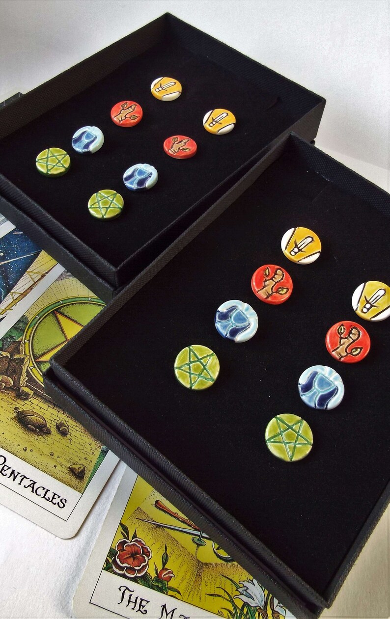 May include: Two black velvet boxes with a variety of colorful round ceramic tokens. The tokens feature different symbols and designs, including pentagrams, swords, and hands. The boxes are open and the tokens are displayed on a black velvet surface.