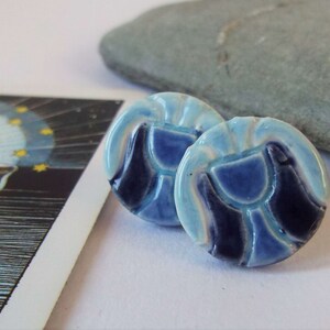 May include: A pair of blue and white ceramic earrings with a geometric design. The earrings are round and have a raised design of a cup or chalice.
