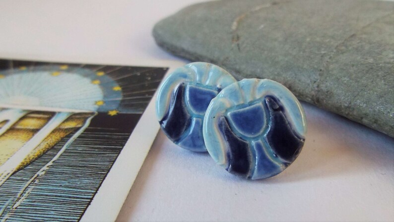 May include: A pair of blue and white ceramic earrings with a geometric design. The earrings are round and have a raised design of a cup or chalice.