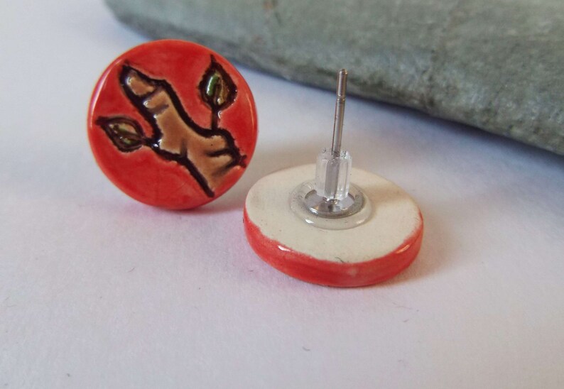May include: A pair of red ceramic earrings with a brown and green twig design. The earrings have silver posts and backs.