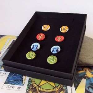 May include: A black box with a velvet interior displays six ceramic round tokens. The tokens feature various symbols, including a sword, a hand, a cup, and a pentagram. The tokens are arranged in two rows of three.