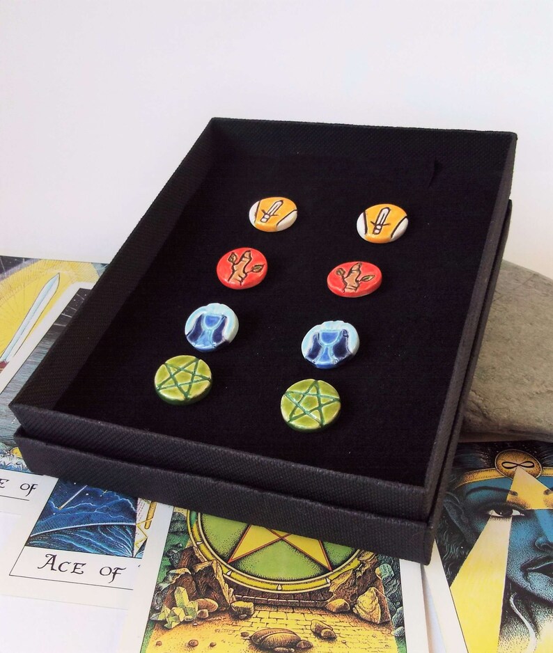 May include: A black box with a velvet interior displays six ceramic round tokens. The tokens feature various symbols, including a sword, a hand, a cup, and a pentagram. The tokens are arranged in two rows of three.