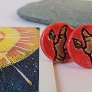 May include: A pair of red ceramic earrings with a hand and leaves design. The hand is brown and the leaves are green.